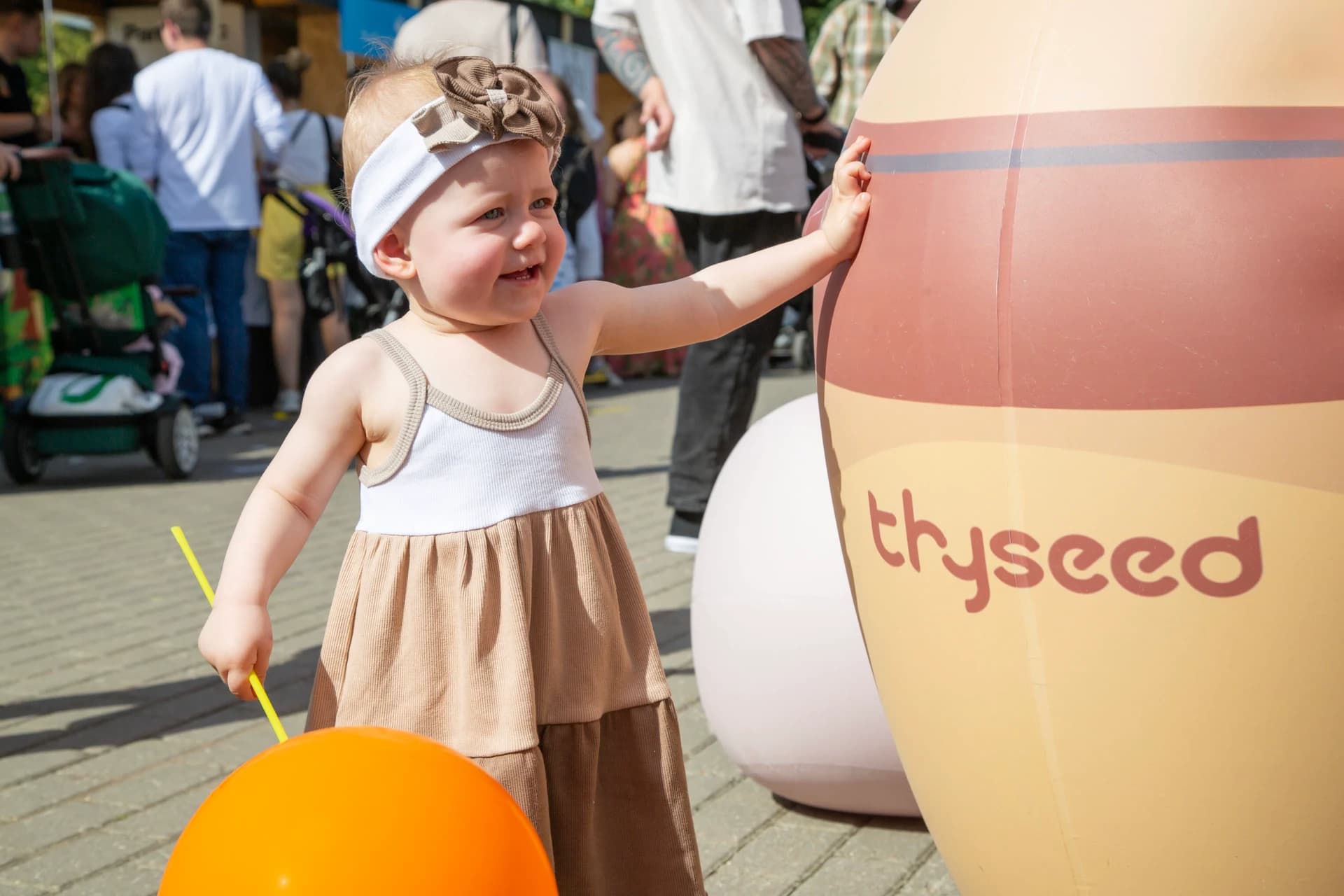 Девочка с оранжевым шариком трогает большую бутылочку Thyseed на празднике